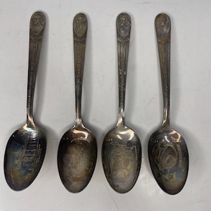 May include: Four vintage silver spoons. Each spoon has a detailed design on the bowl and handle. The handles feature a bust at the top. The bowls have different engraved images and text. A collectible set of spoons.