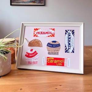 May include: A framed print featuring illustrations of Tunnock's confectionery products, including a Caramel Wafer Biscuit, a Caramel Log, a dark chocolate teacake, and a milk chocolate teacake, all arranged on a light pink background.
