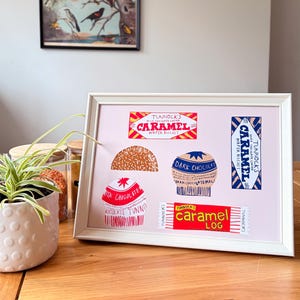 May include: A framed print featuring colorful illustrations of Tunnock's confectionery products, including a Caramel Wafer Biscuit, a Caramel Log, and various chocolate-coated treats, all displayed on a light pink background within a white frame.