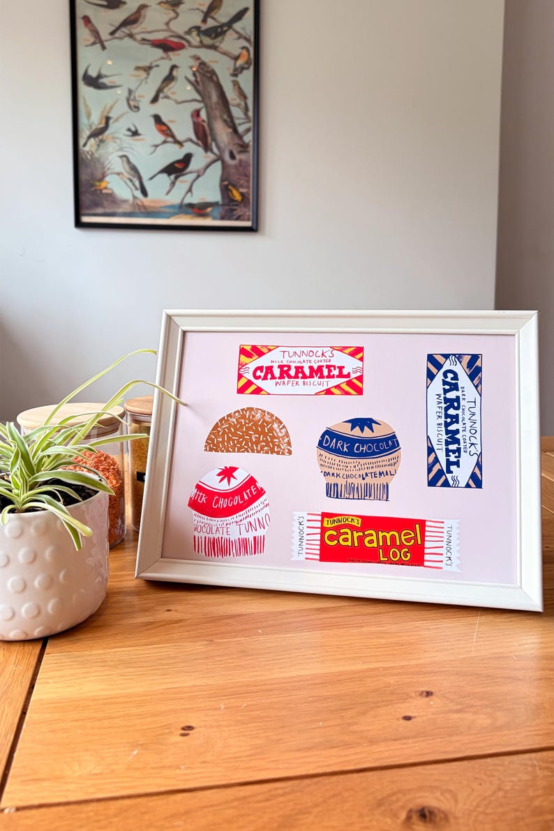 May include: A framed print featuring colorful illustrations of Tunnock's confectionery products, including a Caramel Wafer Biscuit, a Caramel Log, and various chocolate-coated treats, all displayed on a light pink background within a white frame.