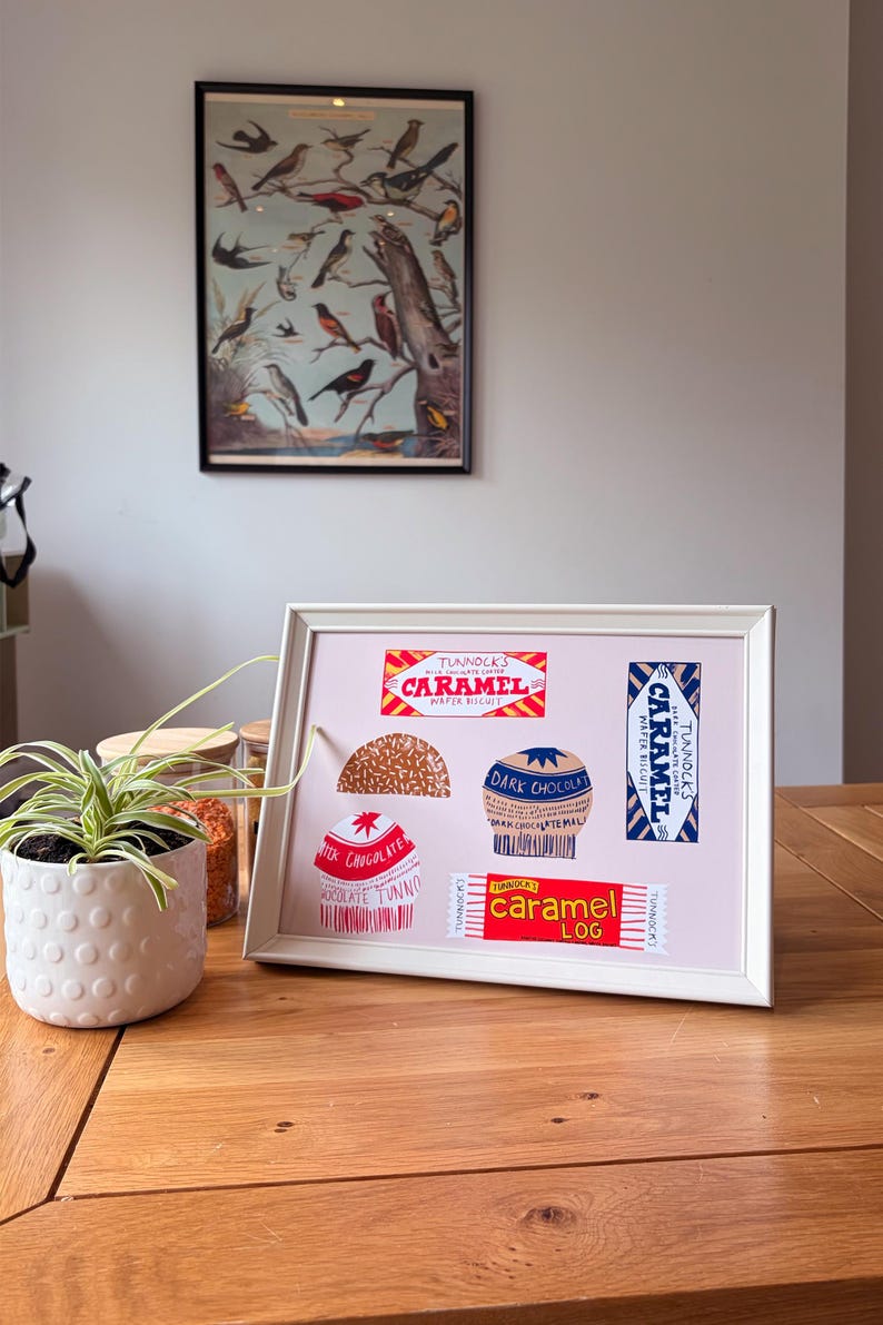 May include: A framed print featuring illustrations of Tunnock's confectionery products, including a Caramel Wafer Biscuit, a Caramel Log, a dark chocolate teacake, and a milk chocolate teacake, all arranged on a light pink background.