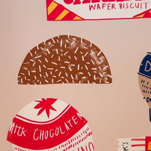 May include: A graphic print featuring a Tunnock's Milk Chocolate Coated Caramel Wafer Biscuit label, a brown semi-circle with white sprinkles, and a red and white Tunnock's Milk Chocolate wrapper design with text and vertical red stripes.