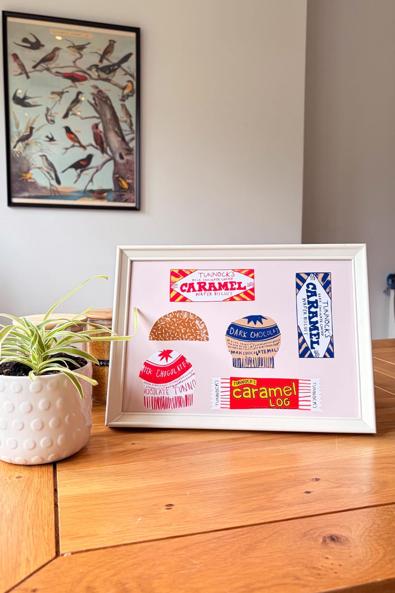 May include: A framed print on a light pink background featuring illustrations of Tunnock's confectionery products, including a Caramel Wafer Biscuit, a Caramel Log, and various chocolate-covered treats with red, blue, and white branding.