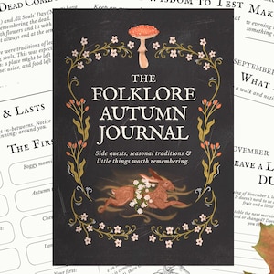 Może przedstawiać: Czarna okładka dziennika z białym napisem The Folklore Autumn Journal, zawierająca centralną ilustrację brązowego zająca otoczonego wieńcem kwiatowym i grzybem u góry, na tle zadrukowanych stron z sezonowymi aktywnościami.