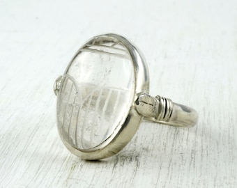 May include: A silver ring featuring an oval, clear stone with an etched design. The ring has a simple band and decorative accents on the sides of the stone. The ring is set against a white background.