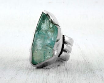 May include: A silver ring with a large, light blue, raw aquamarine gemstone. The ring has a thick band with a textured design.