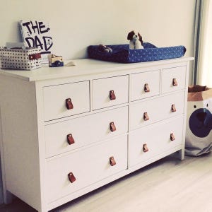 May include: White dresser with eight drawers and brown leather pulls. The dresser has a changing pad on top with a blue and white patterned fabric. A stuffed dog toy sits on the changing pad.