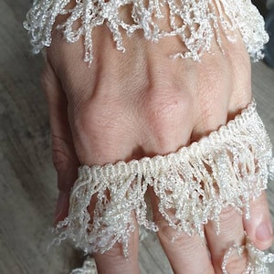 May include: Two white crocheted trim pieces with fringe. The trim is shown on a hand.