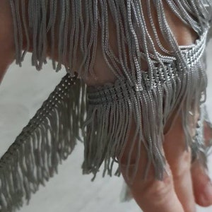 May include: A close-up of silver fringe trim with a woven texture. The fringe is soft and delicate, with a subtle sheen.