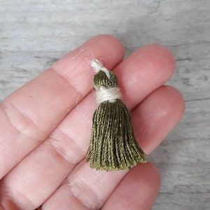 Könnte beinhalten: Ein kleiner grüner Quaste mit einer weißen Schnur. Die Quaste ist etwa 2,5 cm lang und aus weichen, seidenartigen Fäden gefertigt.