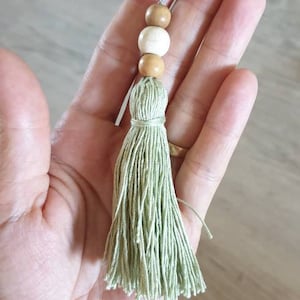 May include: A handmade tassel with a light green fringe and wooden beads. The tassel is made of green thread and has three wooden beads in shades of brown and white. The tassel is held up by a metal loop.