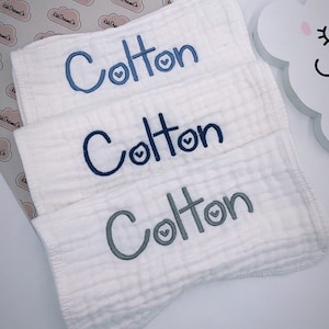 May include: Three white cotton burp cloths with the name "Colton" embroidered in blue and gray thread. Each name has a small heart embroidered next to it.
