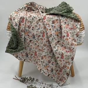 May include: A baby blanket with a whimsical forest print featuring mushrooms, insects, and whimsical figures in shades of beige, orange, and green. The blanket has a soft, textured green backing. The blanket is draped over a small wooden stool.