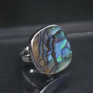 May include: A silver ring with a square abalone shell set in the center. The shell has iridescent blue, green, and brown colors.