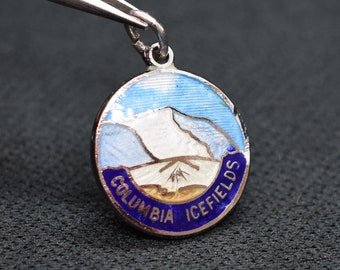 May include: A silver charm with a blue and white enamel design depicting a mountain range. The charm is inscribed with the words "Columbia Icefields".
