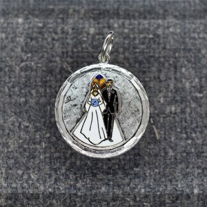 May include: Silver charm with a white background and a bride and groom in black and white with colored accents. The bride is wearing a white dress and a blue flower. The groom is wearing a black suit. The charm is round and has a textured surface.