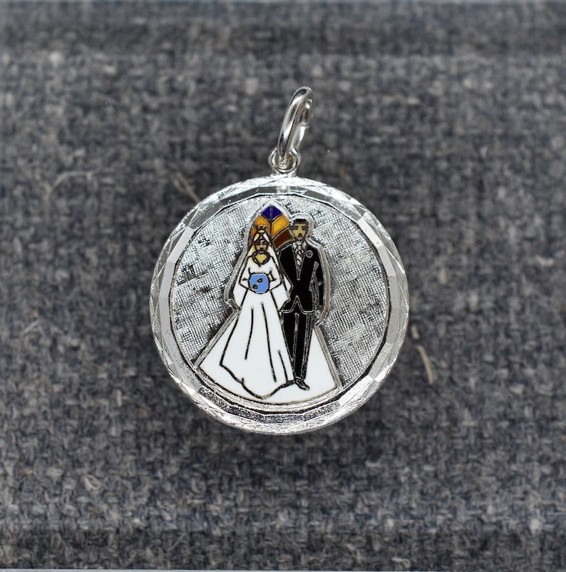 May include: Silver charm with a white background and a bride and groom in black and white with colored accents. The bride is wearing a white dress and a blue flower. The groom is wearing a black suit. The charm is round and has a textured surface.