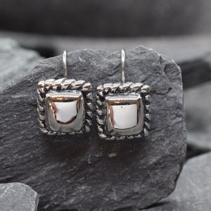 May include: A pair of silver dangle earrings with a square design. The earrings have a rope-like border and a smooth, reflective surface.