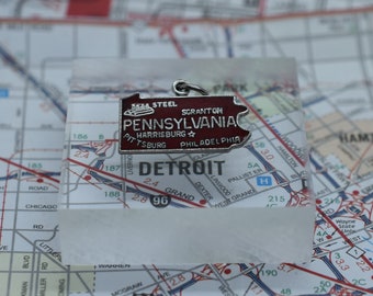 May include: A silver charm with a red enamel state of Pennsylvania charm. The charm has the words "Gas Steel Scranton Pennsylvania Harrisburg Pittsburgh Philadelphia" on it. The charm is sitting on a map of Detroit.