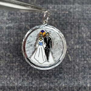 May include: Silver charm with a white enamel bride and groom in a wedding scene. The charm has a textured silver background and a loop for attaching to a chain.