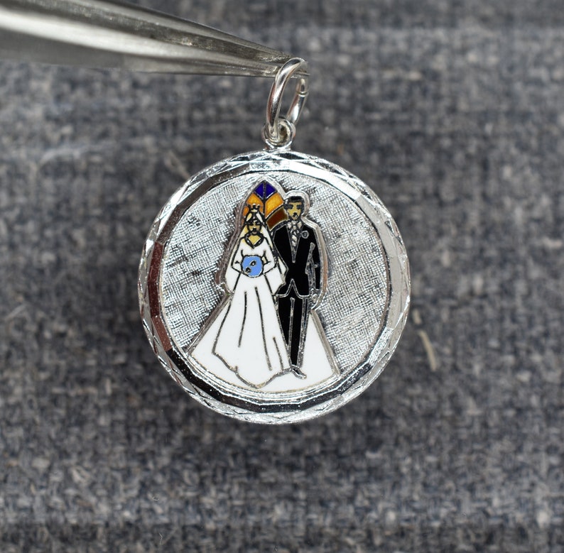 May include: Silver charm with a white enamel bride and groom in a wedding scene. The charm has a textured silver background and a loop for attaching to a chain.