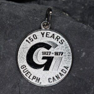 May include: A silver pendant with a circular design. The pendant commemorates "150 YEARS" with the years "1827-1977" and the text "GUELPH, CANADA". The pendant has a loop for attaching to a chain.