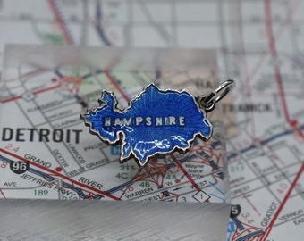 May include: A silver charm in the shape of the state of New Hampshire. The charm is blue with a white outline and the word "HAMPSHIRE" in white letters. The charm is on a map of Detroit, Michigan.
