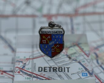 May include: A silver charm with a blue and red enamel coat of arms. The coat of arms features a shield with a red cross and a white and blue design. The charm is inscribed with the text "Berchtesgaden". The charm is sitting on a map of Detroit.