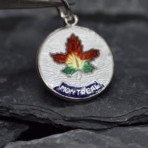 May include: A silver charm with a white enamel background featuring a colorful maple leaf and the word "MONTREAL" in blue enamel.
