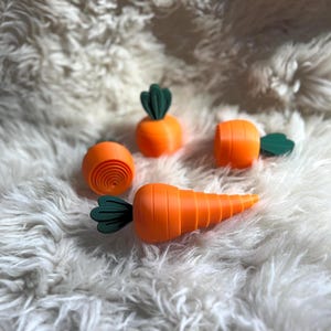 May include: An assortment of orange carrot-shaped objects with green tops, arranged on a fluffy white surface. The carrots vary in size and shape, with some appearing as slices and others as whole carrots. The objects are likely decorative or toy items.