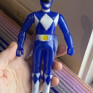 May include: A blue action figure of a Power Ranger, featuring a white diamond on the chest and white accents on the boots. The figure has a yellow belt buckle and a helmet with a black visor. The toy is held in a hand.