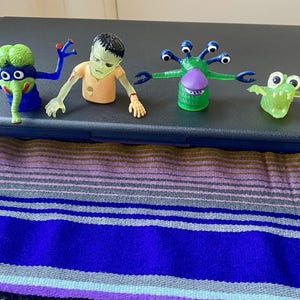 May include: Four colorful monster figurines. The first is blue and green with a trunk, the second is a zombie, the third is green with multiple eyes, and the fourth is a green frog-like creature.