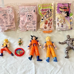 May include: A collection of Dragon Ball Z and Powerpuff Girls toys, including action figures and sealed bags. The figures are in various poses and colors, with orange, yellow, and silver figures. The bags feature character illustrations.