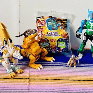 May include: A set of four action figures, including a blue and green creature with a large head, a yellow and orange dinosaur, a white and gold creature with a long tail, and a small silver robot. The figures are on a striped rug. The packaging for a Fox Kids Mini-Viewer is visible in the background.