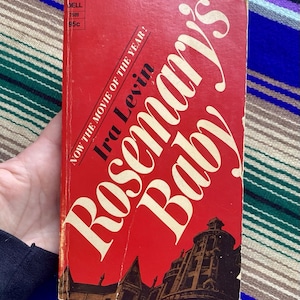 May include: A vintage paperback book titled "Rosemary's Baby" by Ira Levin. The cover is predominantly red with white lettering and a dark illustration of a building. The text "NOW THE MOVIE OF THE YEAR!" is also present.