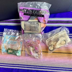 May include: Several sealed toy sets, including a motorcycle, a car, and other small toys. The packaging features the text "X-STUNT SPORT SETS" and warnings about small parts and age recommendations. The toys are on a striped surface.