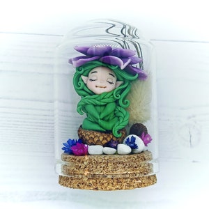 Puede incluir: Una pequeña figura de hada verde con flores moradas y hojas verdes dentro de un frasco de vidrio transparente con tapa de corcho.