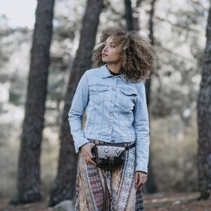 May include: A woman wearing a light blue denim jacket and a patterned skirt with a brown leather fanny pack with a snake skin pattern.