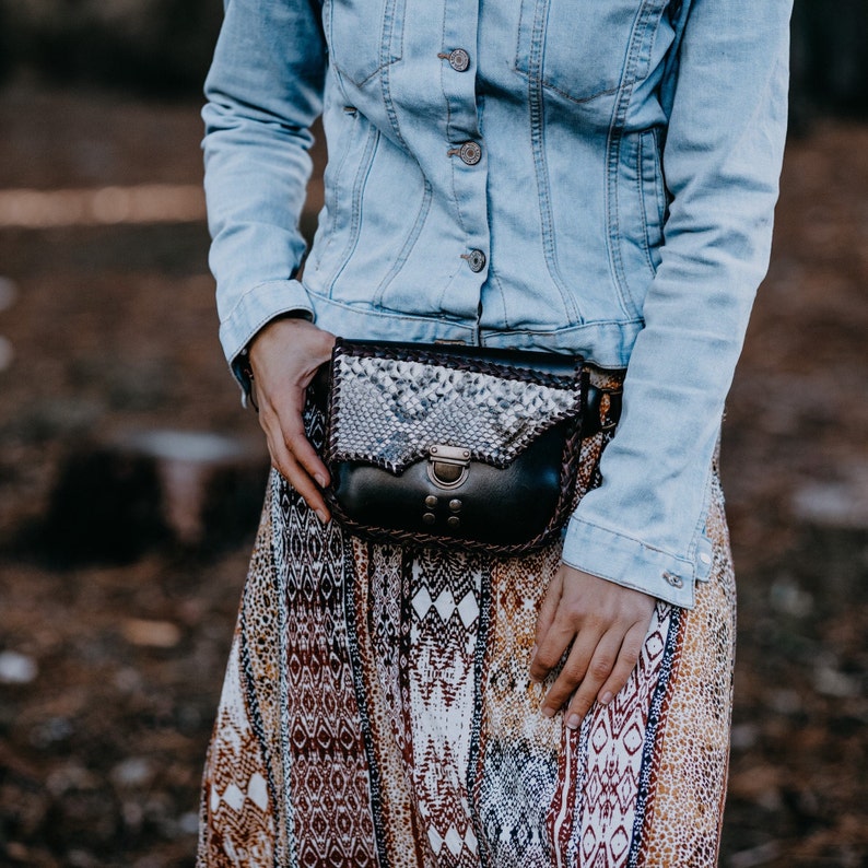 May include: A black and white snakeskin patterned leather fanny pack with a brown leather strap and a silver buckle. The fanny pack is worn over a denim jacket and a patterned skirt.