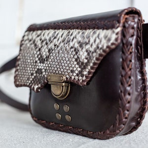 May include: A brown leather crossbody bag with a snakeskin flap and a brass buckle closure. The bag has a braided leather trim and decorative studs.