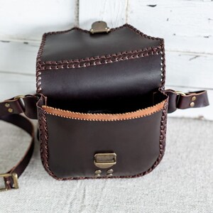 May include: A brown leather bag with a flap closure and a long strap. The bag has a braided design around the edges and a brass buckle on the flap.