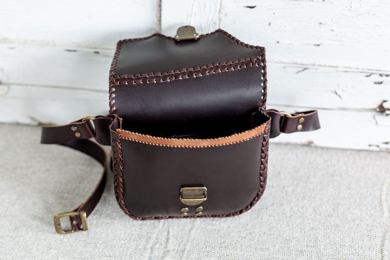 May include: A brown leather bag with a flap closure and a long strap. The bag has a braided design around the edges and a brass buckle on the flap.