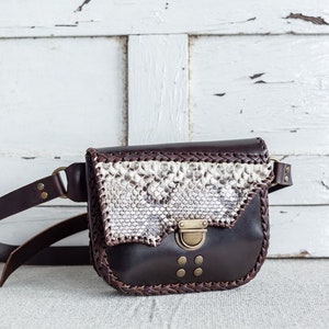 May include: A brown leather fanny pack with a snakeskin pattern flap and a brass buckle closure. The bag has a braided leather trim and a long adjustable strap.