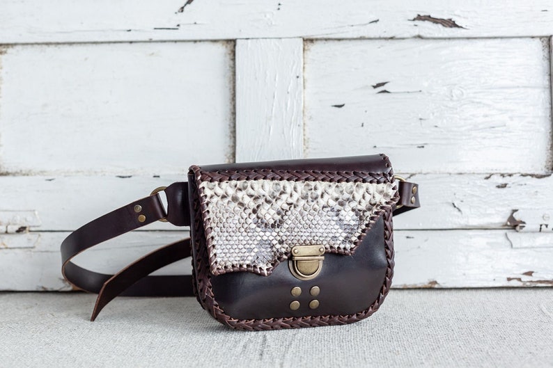 May include: A brown leather fanny pack with a snakeskin pattern flap and a brass buckle closure. The bag has a braided leather trim and a long adjustable strap.