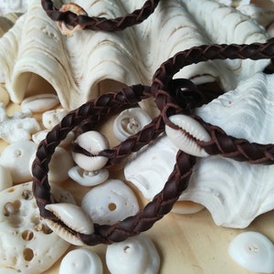 May include: Two brown leather bracelets with white cowrie shells woven into the braid. The bracelets are displayed on a white seashell and surrounded by other white seashells.