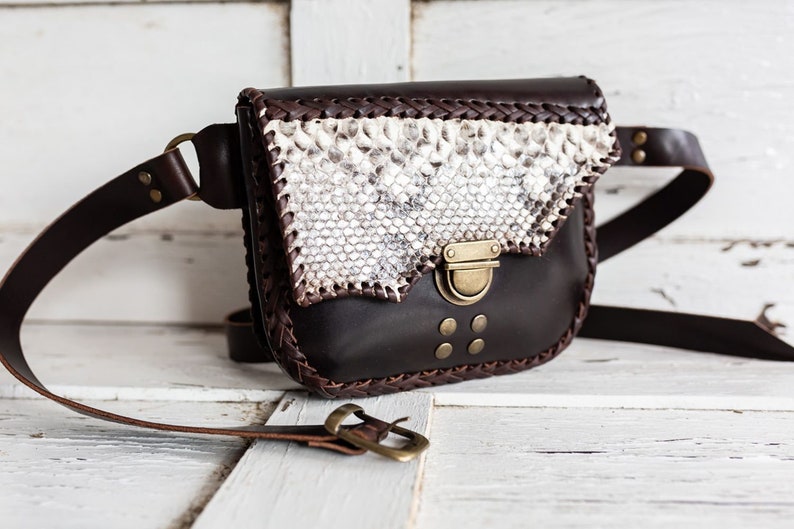 May include: A brown leather fanny pack with a snakeskin flap and a brass buckle. The bag has a braided leather trim and a long adjustable strap.