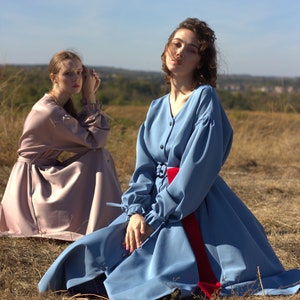 May include: Two women wearing long dresses pose outdoors. The woman in the foreground is wearing a light blue dress with a red sash. The woman in the background is wearing a light pink dress. Both women are sitting in a field of dry grass.