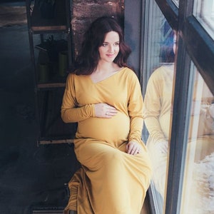 May include: A pregnant person in a long, mustard yellow dress sits by a window, looking out at the snowy landscape.