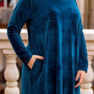 May include: A teal blue velvet dress with long sleeves and a loose fit. The dress has a round neckline and two pockets on the sides.