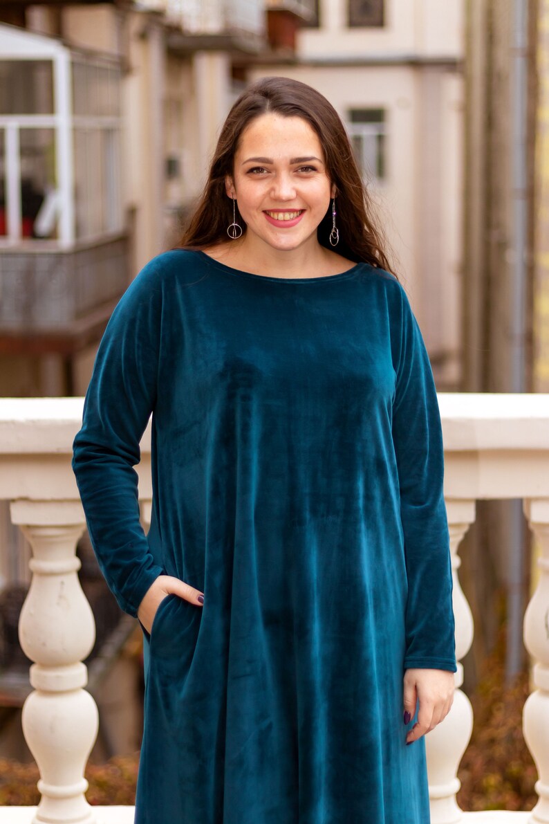 May include: A woman wearing a teal blue velvet dress with long sleeves and a pocket on the left side. She is standing in front of a white railing.